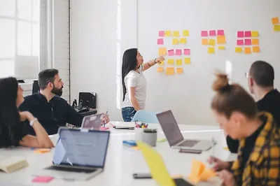 A consultant points to sticky notes on a wall while colleagues around a table listen